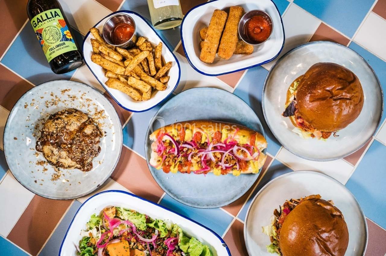 Assortiment de plats comprenant burgers, hot-dog, salade, frites, mozzarella sticks, bière et vin sur une table colorée.