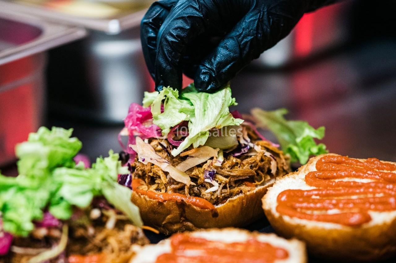 Un cuisinier garnit un burger avec de la laitue et du chou sur une base de viande effilochée et de sauce.