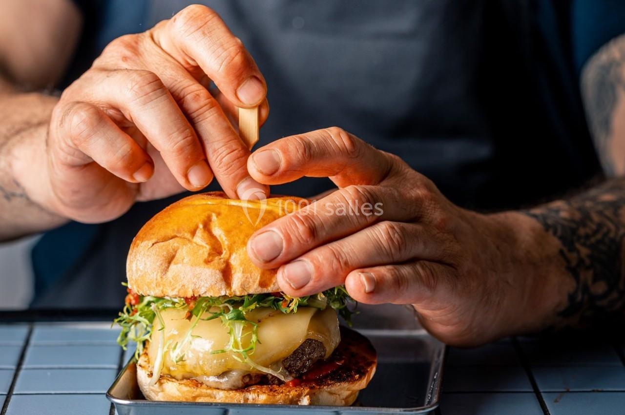 Un cuisinier assemble un burger garni de fromage fondu, de jeunes pousses et de sauce, posé sur un plateau métallique.