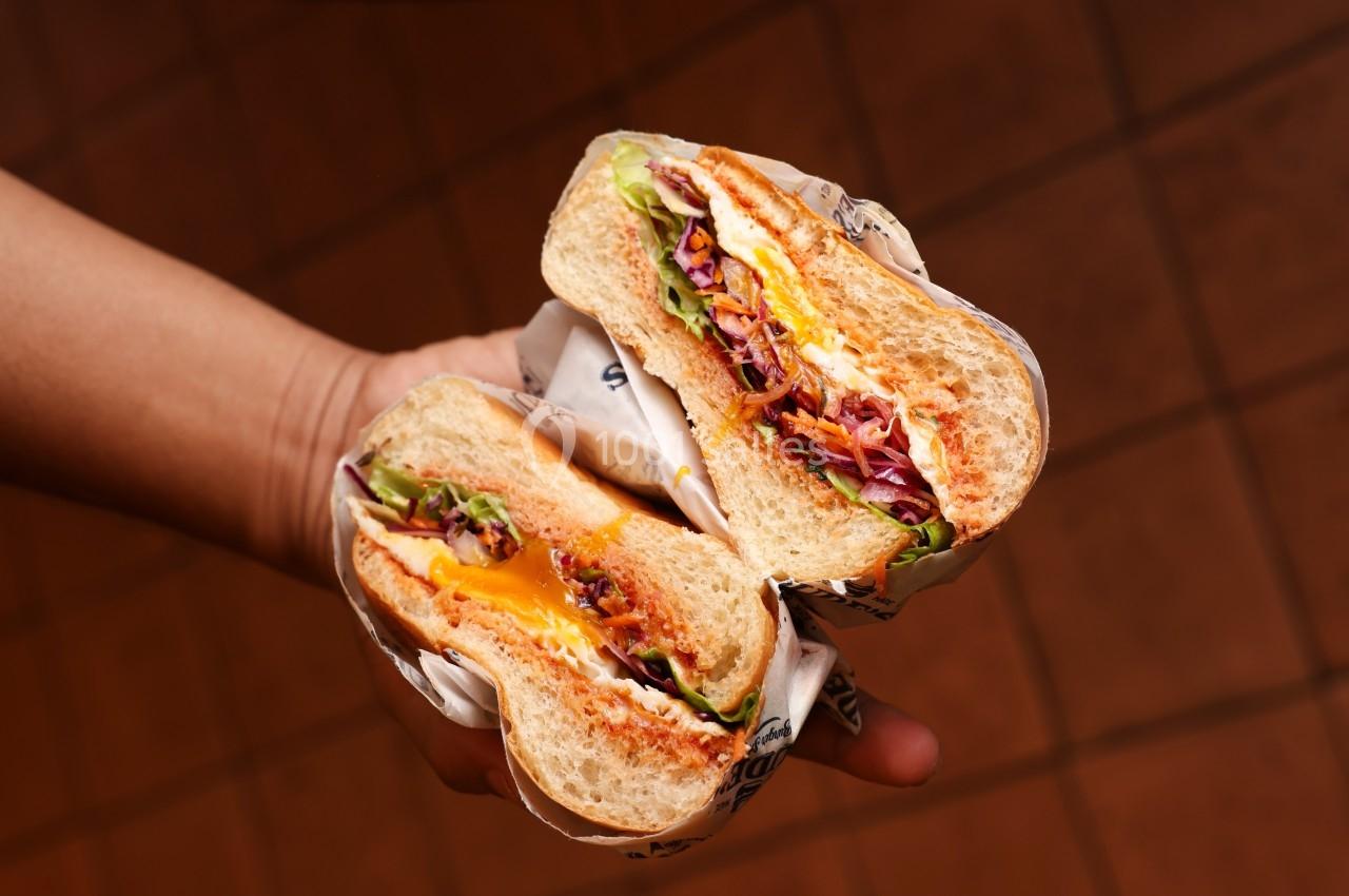 Deux moitiés de sandwich garnies de légumes, fromage fondu et sauce, tenues dans une main sur un fond carrelé.