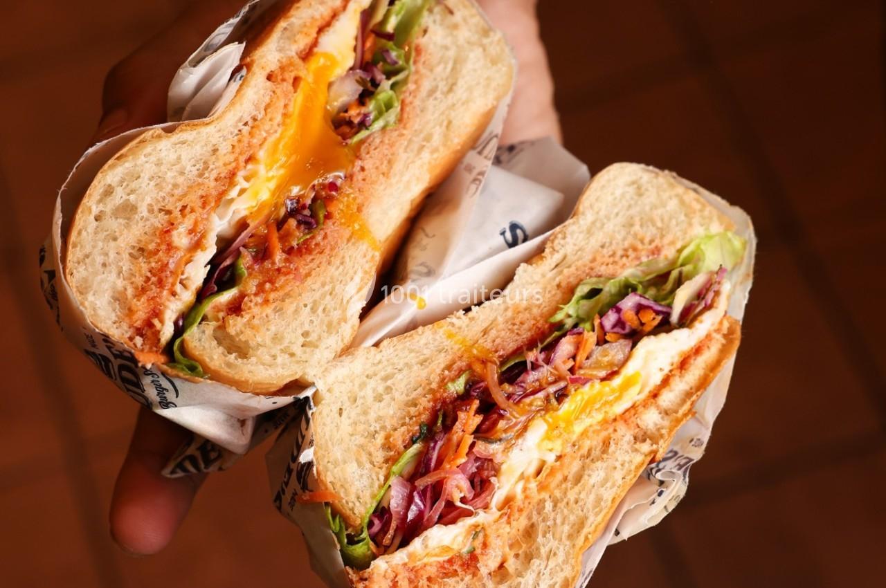 Sandwich coupé en deux, garni de légumes, fromage fondu et sauce, tenu dans une main sur un fond flou.