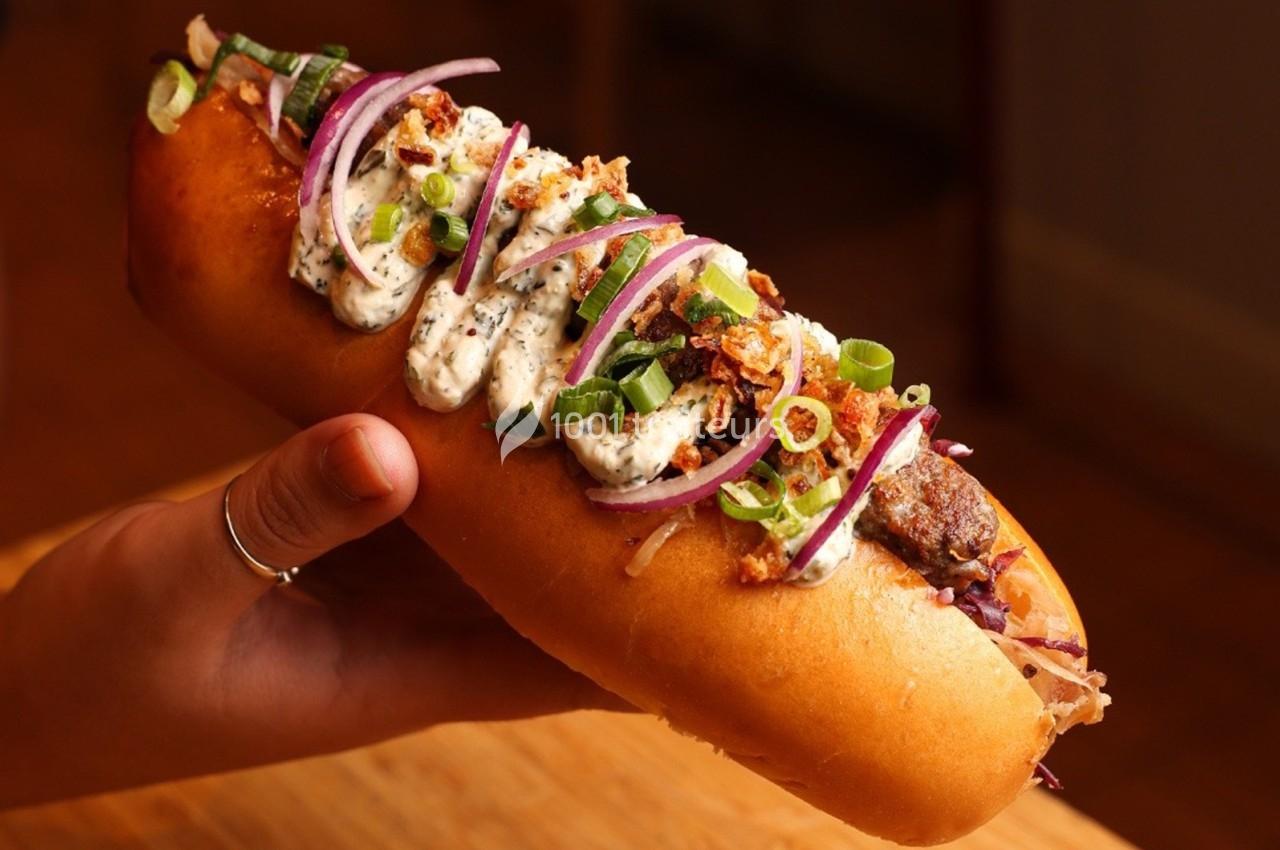 Hot-dog garni de crème, oignons rouges, ciboulette et éclats croustillants, tenu par une main.