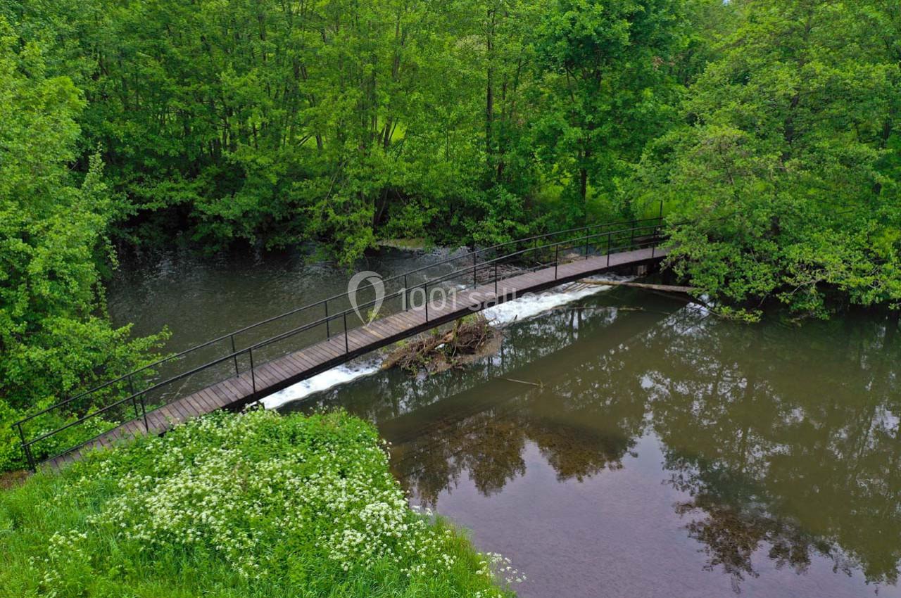 Passerelle en bois avec rambardes traversant une rivière entourée de végétation dense et arbres verdoyants.