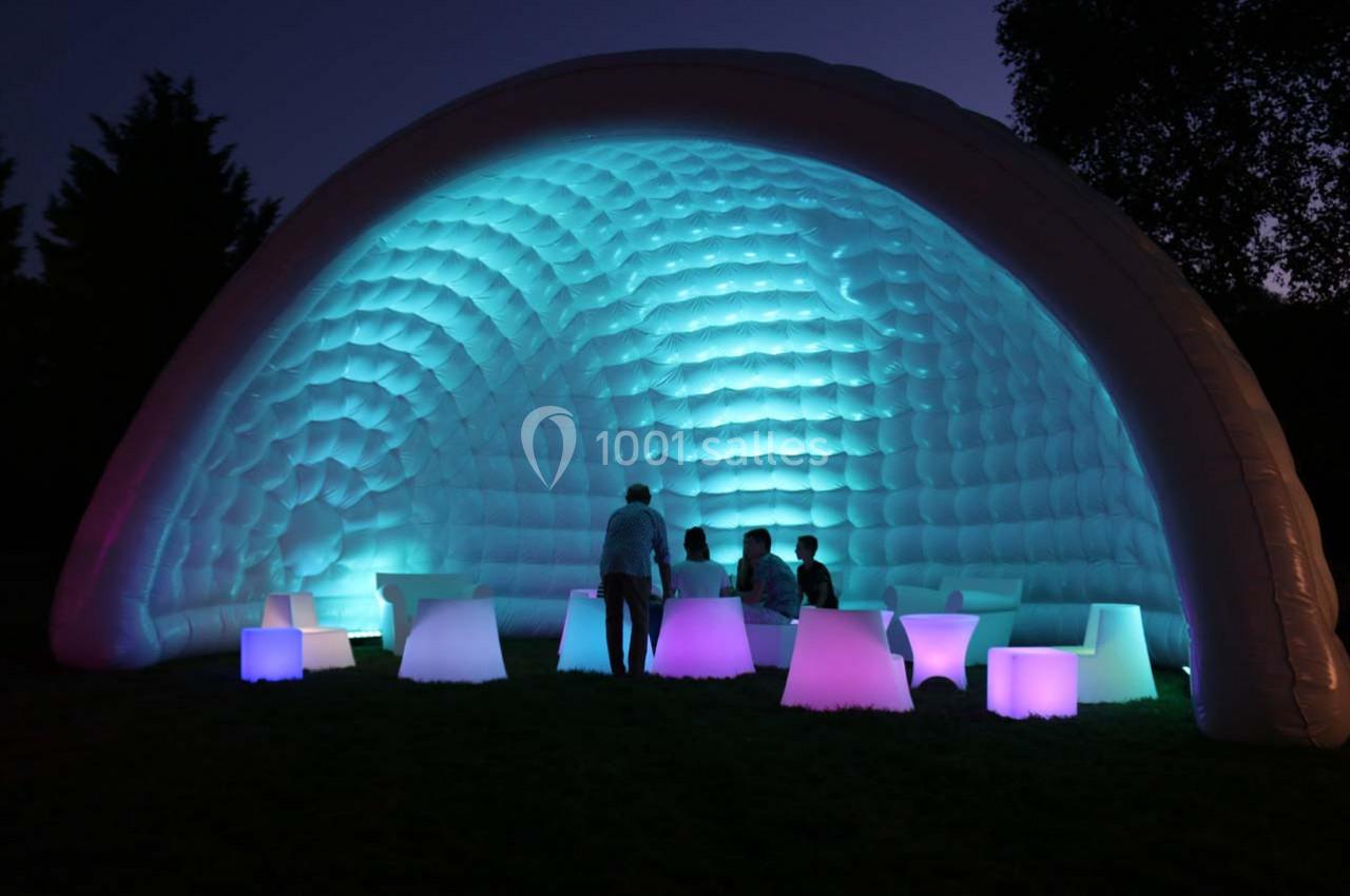 Structure gonflable éclairée en bleu et violet, avec des personnes assises autour de tables lumineuses dans l'obscurité.