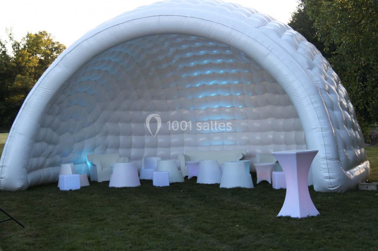 Structure gonflable blanche en forme de dôme avec des sièges et tables lumineux disposés sur une pelouse.