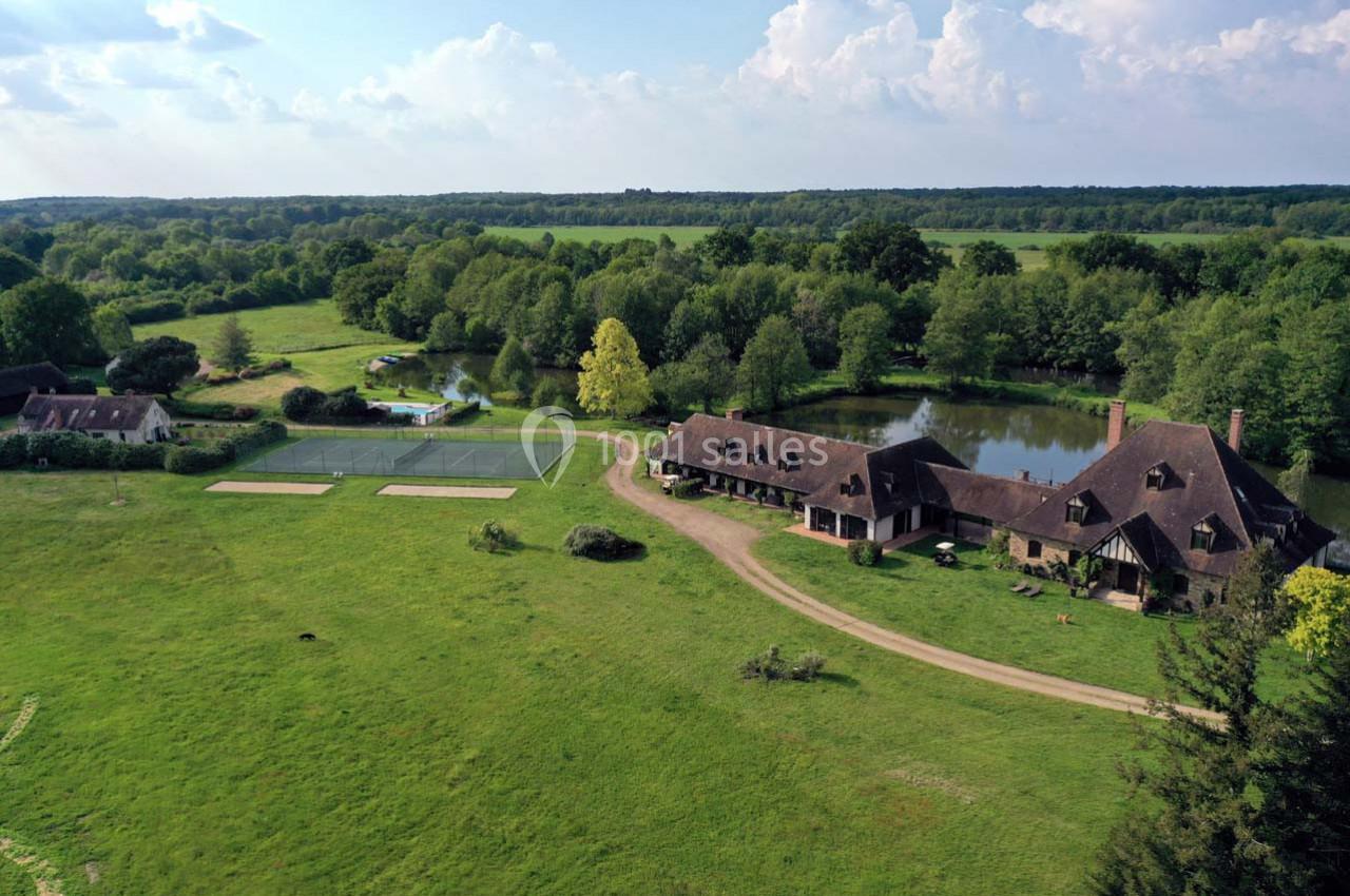 Vue aérienne d'une propriété rurale avec bâtiments, étang, terrains de tennis et prairies entourés de forêt.