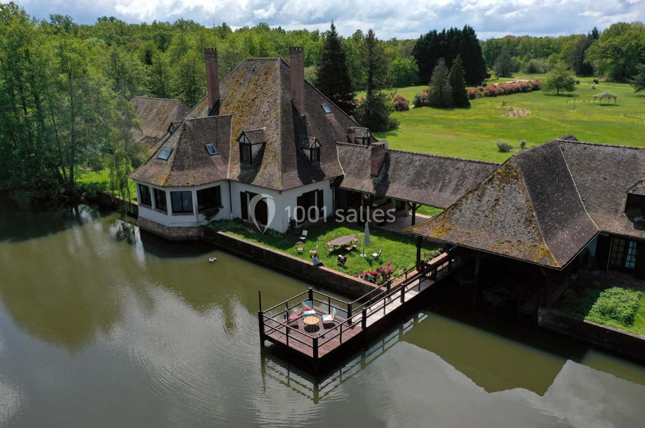 Maison traditionnelle au bord d’un étang, entourée de verdure, avec une terrasse sur pilotis.