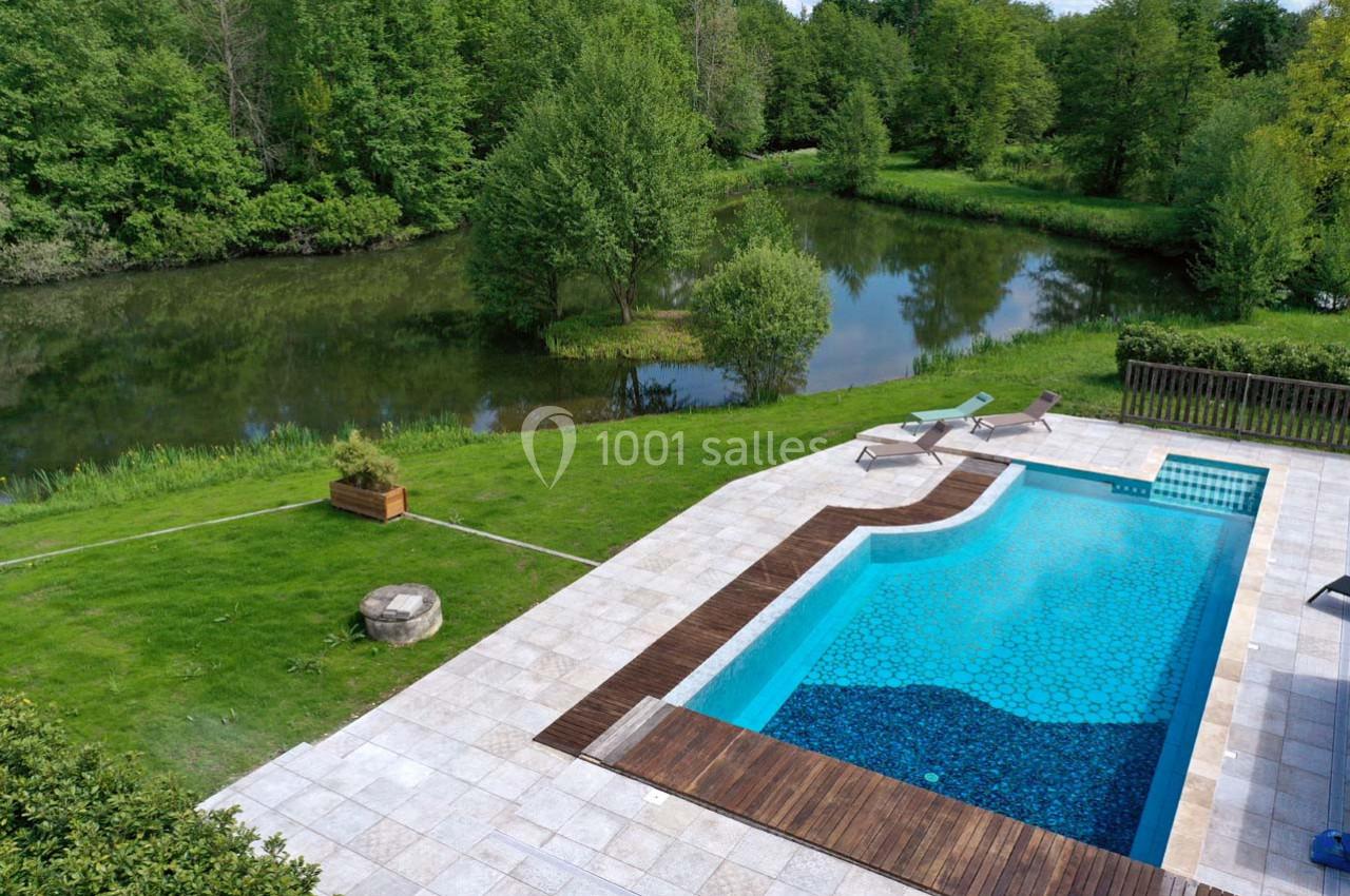 Piscine extérieure avec terrasse en bois, bordée de pelouse, près d'une rivière entourée de végétation.