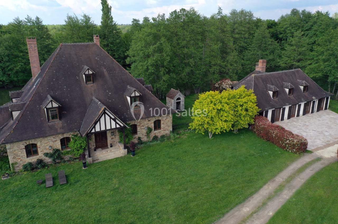 Vue aérienne d'une maison en pierre à colombages avec un toit en pente, un jardin et une dépendance entourés d'arbres.