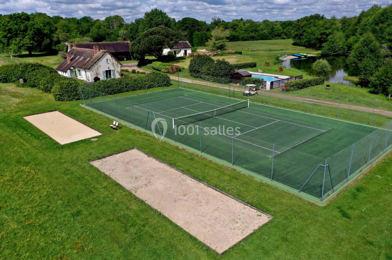 Terrain de tennis extérieur entouré de verdure, avec une maison, un étang et des équipements à proximité.