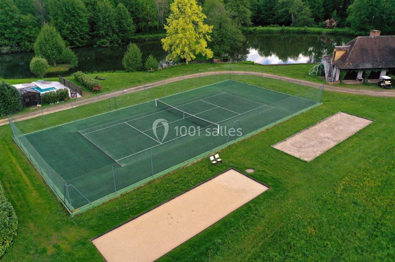 Terrain de tennis extérieur entouré de verdure, avec un étang et une maison en arrière-plan.