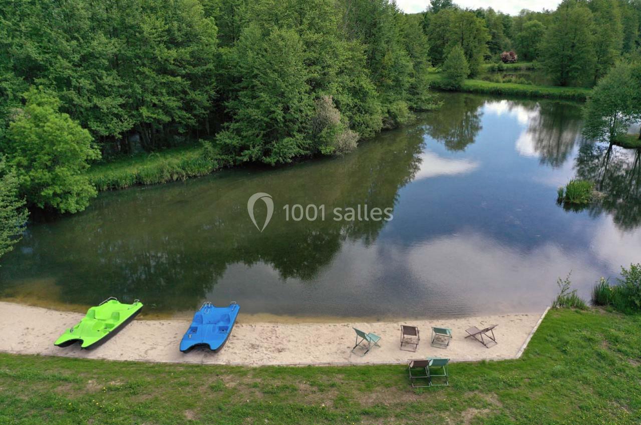 Vue d'un petit lac entouré de forêt avec deux pédalos colorés et des chaises disposées sur une plage de sable.