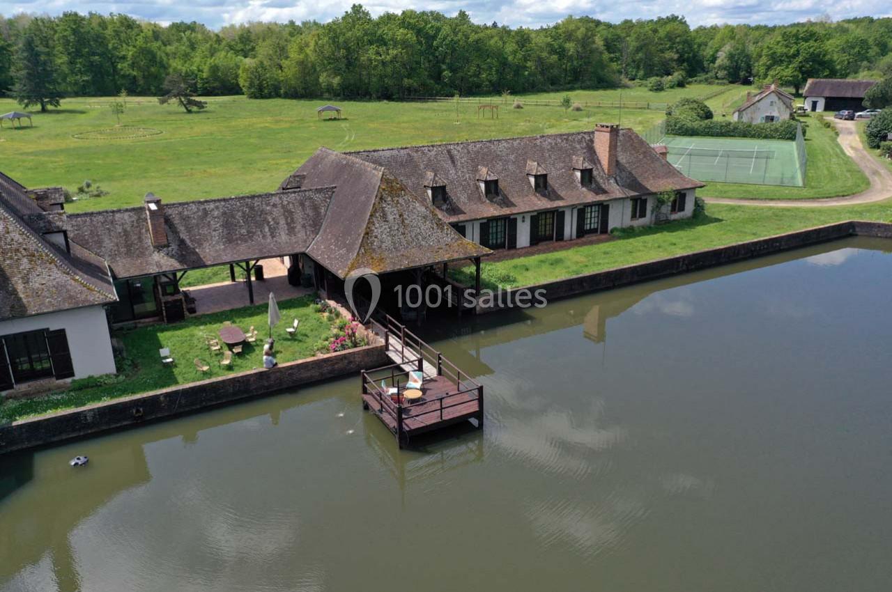 Vue aérienne d'une maison traditionnelle avec toit en chaume, bordée d'un étang et entourée de verdure.