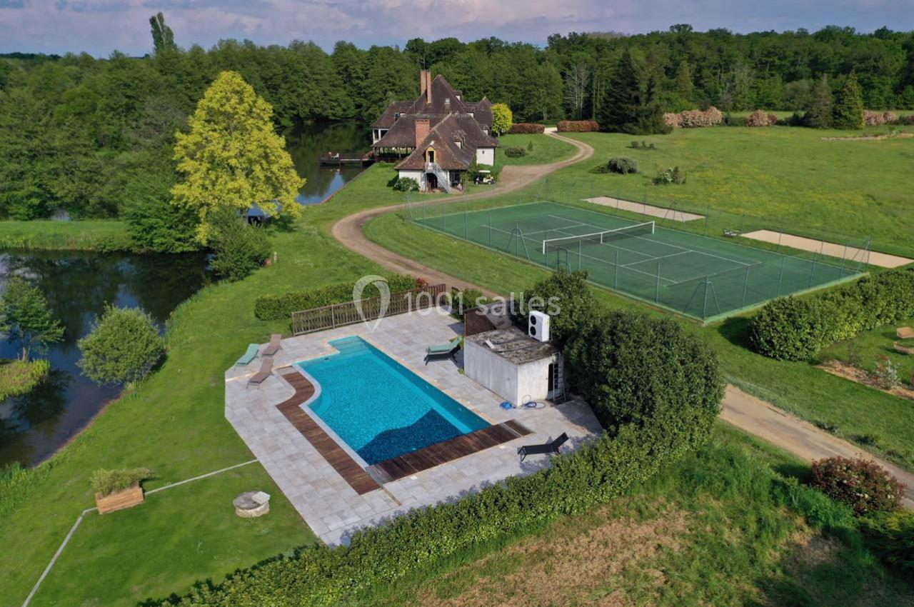 Vue aérienne d'une propriété avec piscine, terrain de tennis, étang et maison entourés de verdure.