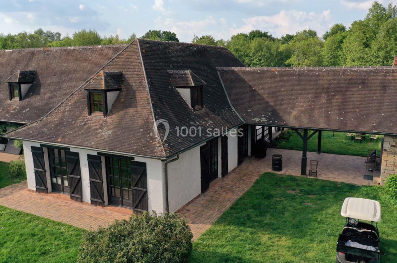 Bâtiment rural avec toit en ardoise, grandes fenêtres et terrasse pavée, entouré de pelouse et d'arbres.