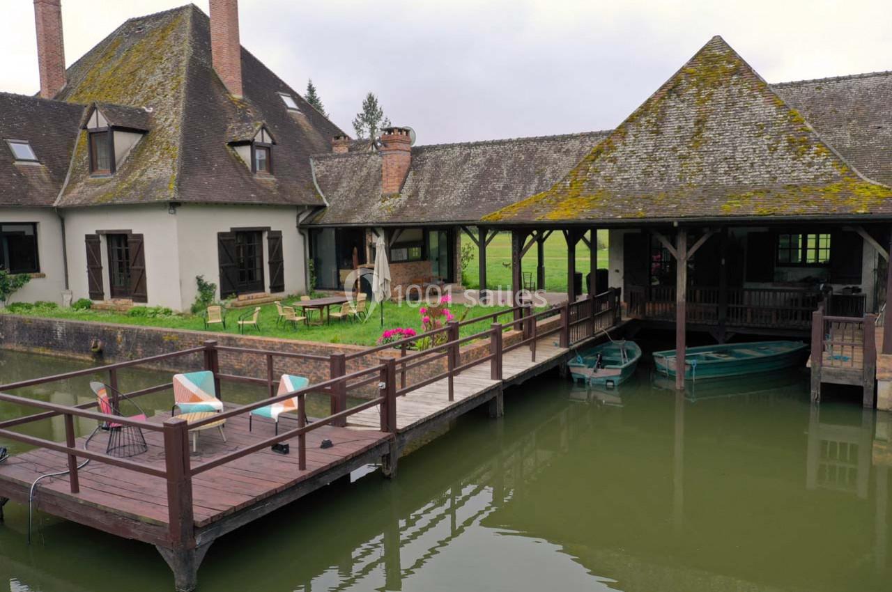 Ponton en bois avec chaises et table, donnant sur un plan d'eau, près d'une maison à toit en ardoise et jardin verdoyant.
