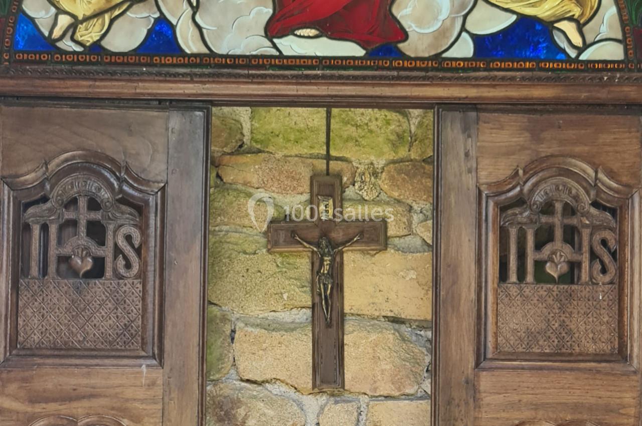 Croix en bois accrochée sur un mur en pierre, encadrée par des portes sculptées et un vitrail coloré au-dessus.