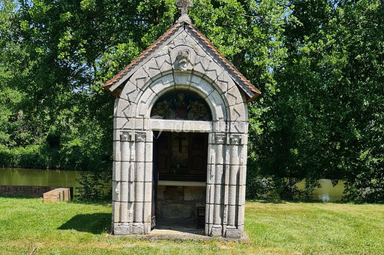 Petite chapelle en pierre avec un toit en tuiles, entourée d'herbe et d'arbres, près d'un plan d'eau.