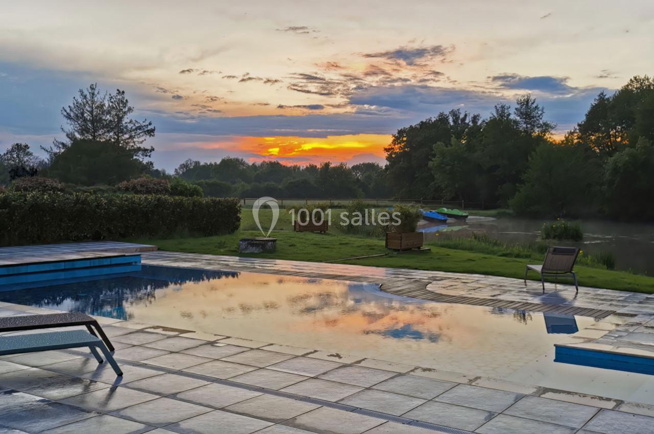 Piscine extérieure avec terrasse en pierre, vue sur un étang et un coucher de soleil coloré derrière des arbres.