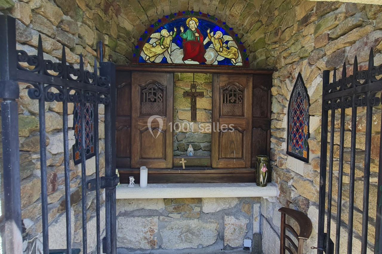 Autel en pierre avec croix en bois, vitrail coloré représentant une scène religieuse et grilles en fer forgé ouvertes.