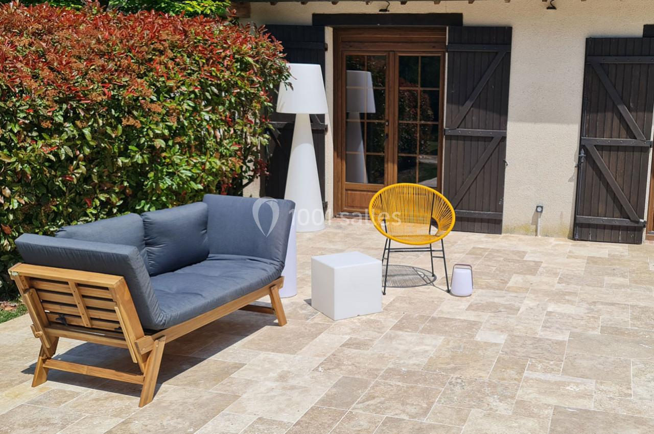 Salon de jardin avec canapé en bois, fauteuil jaune, table basse blanche et haie en arrière-plan sur une terrasse pavée.