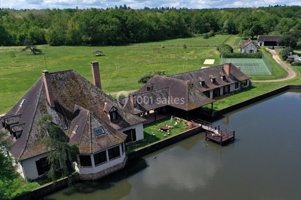 Vue aérienne d'une grande maison en bord d'eau entourée de verdure, avec un terrain et des bâtiments annexes.