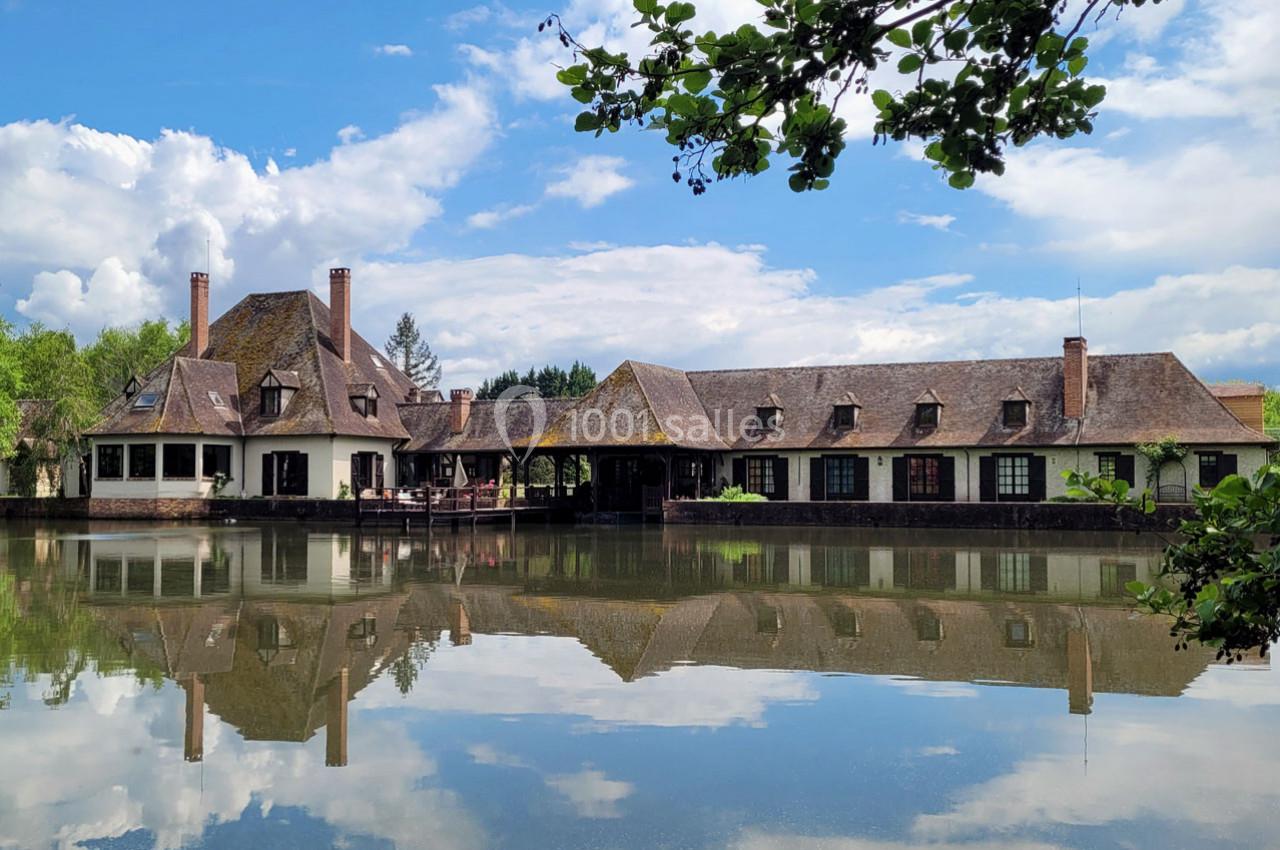 Grande maison au bord d'un étang calme, avec un ciel partiellement nuageux et des arbres en arrière-plan.