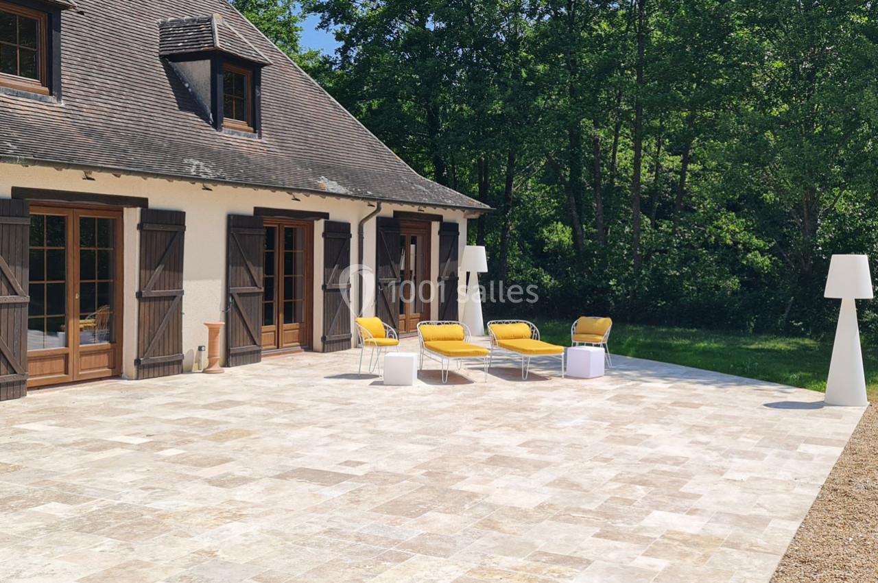 Terrasse en pierre devant une maison avec des volets en bois, mobilier jaune et blanc, entourée de verdure.