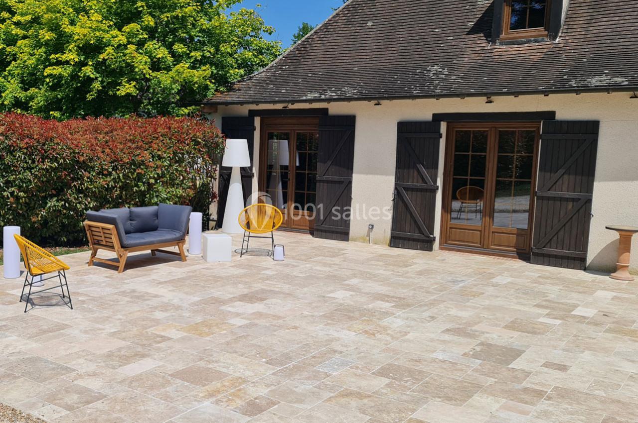 Terrasse en pierre avec mobilier de jardin moderne, devant une maison aux volets en bois et entourée de végétation.