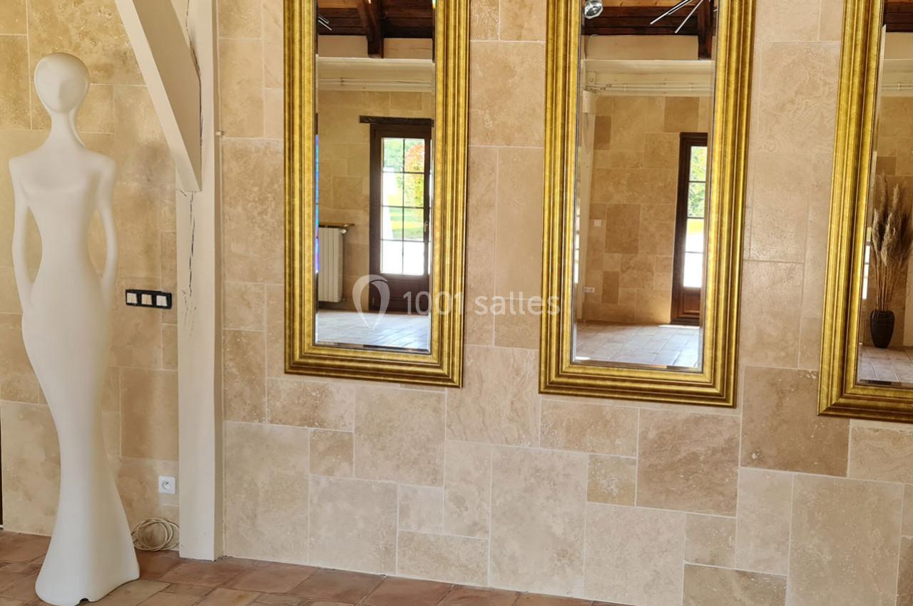 Mannequin blanc stylisé près de trois miroirs dorés accrochés à un mur en pierre beige dans une pièce lumineuse.