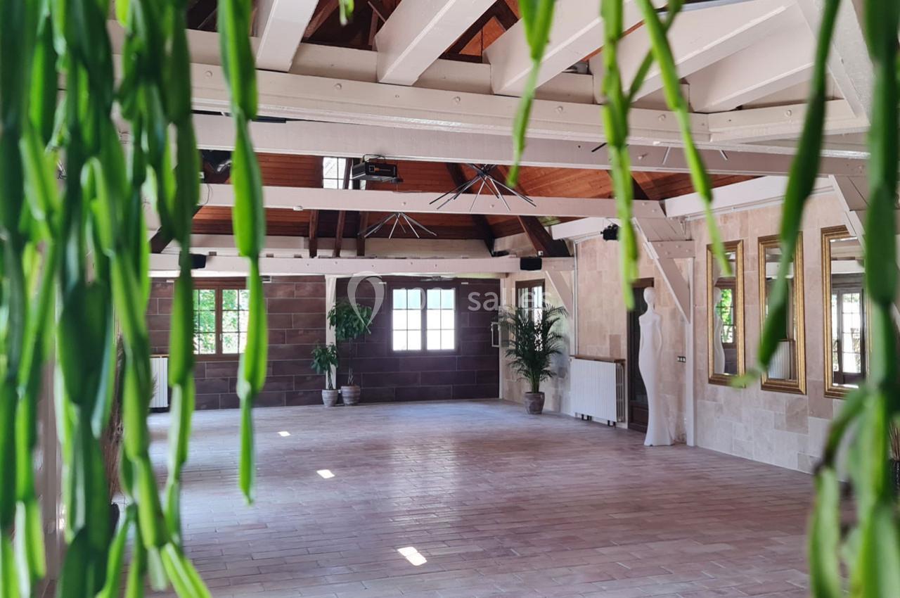 Salle spacieuse avec poutres apparentes, grandes fenêtres, plantes et murs en pierre dans un cadre lumineux.