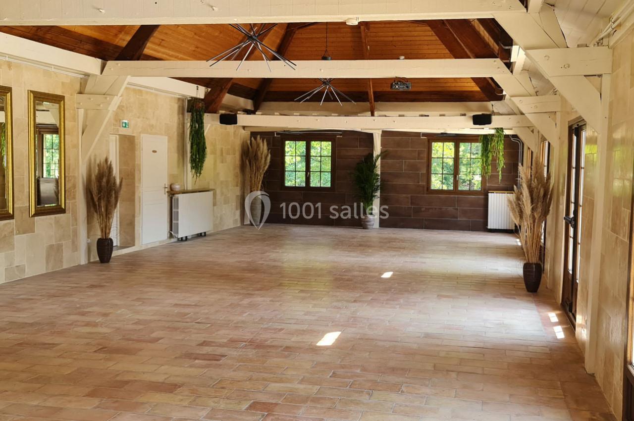 Salle lumineuse avec murs en pierre, poutres apparentes, grandes fenêtres, plantes décoratives et sol en carrelage.