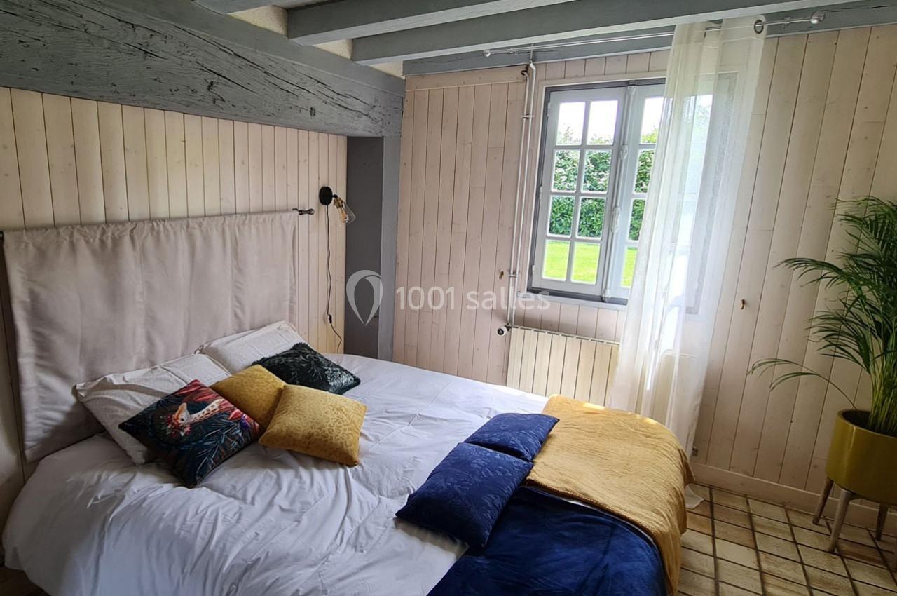 Chambre lumineuse avec lit double, coussins colorés, murs en bois clair, fenêtre donnant sur un jardin et plante en pot.