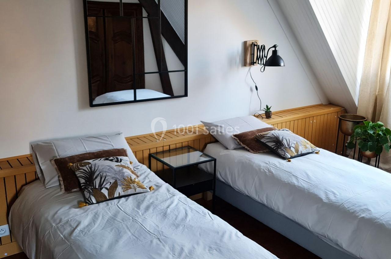 Deux lits simples avec coussins décoratifs dans une chambre lumineuse, équipée d'une lampe murale et d'une plante verte.