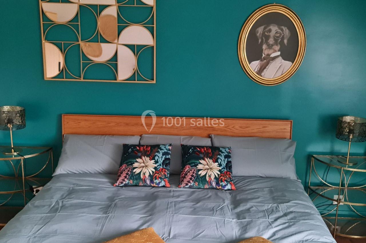 Lit double avec draps bleus, coussins à motifs floraux, mur turquoise décoré d'un tableau et d'un miroir géométrique.