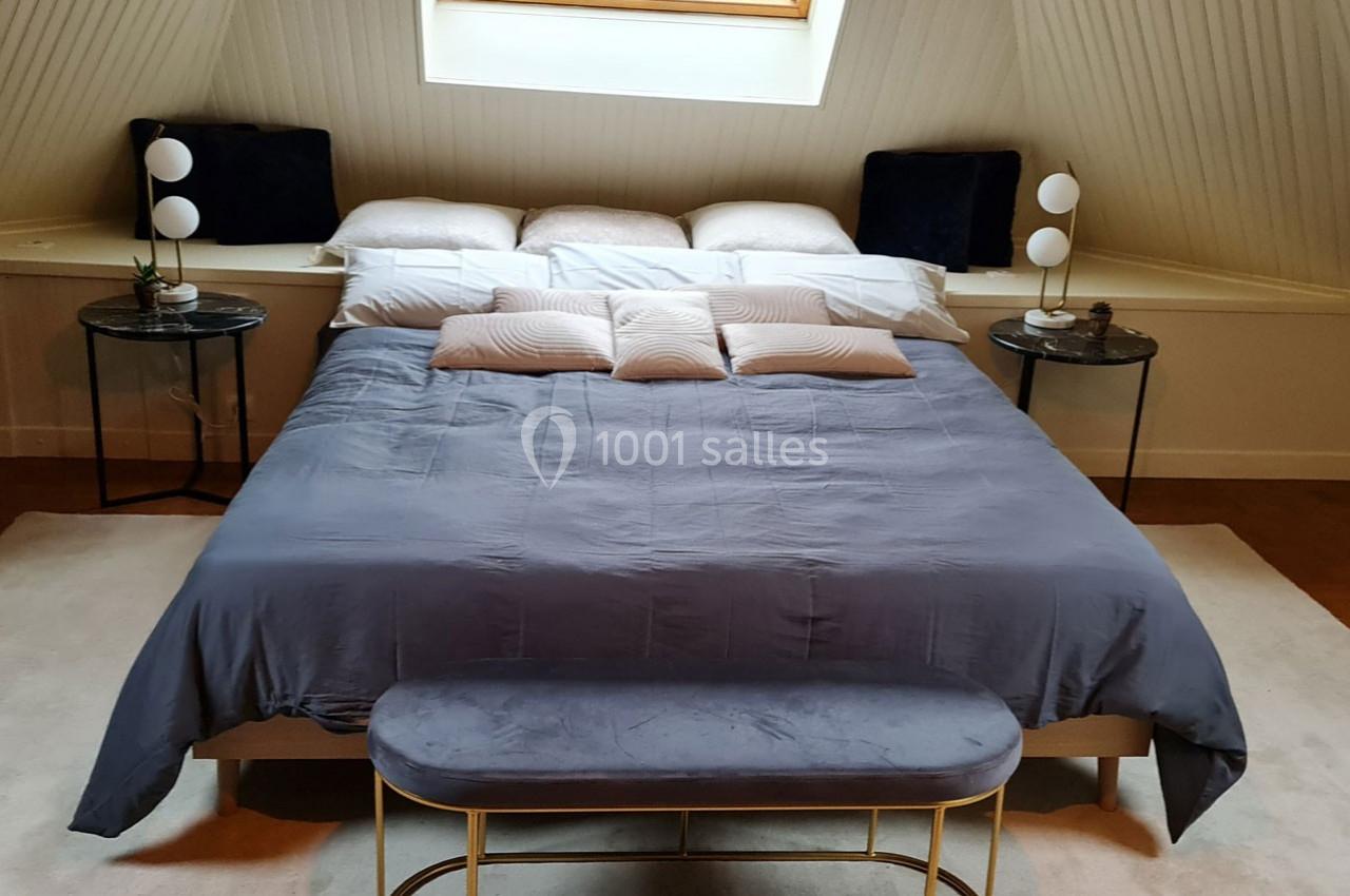 Lit double avec draps gris, coussins décoratifs et tables de chevet surmontées de lampes, dans une chambre mansardée.