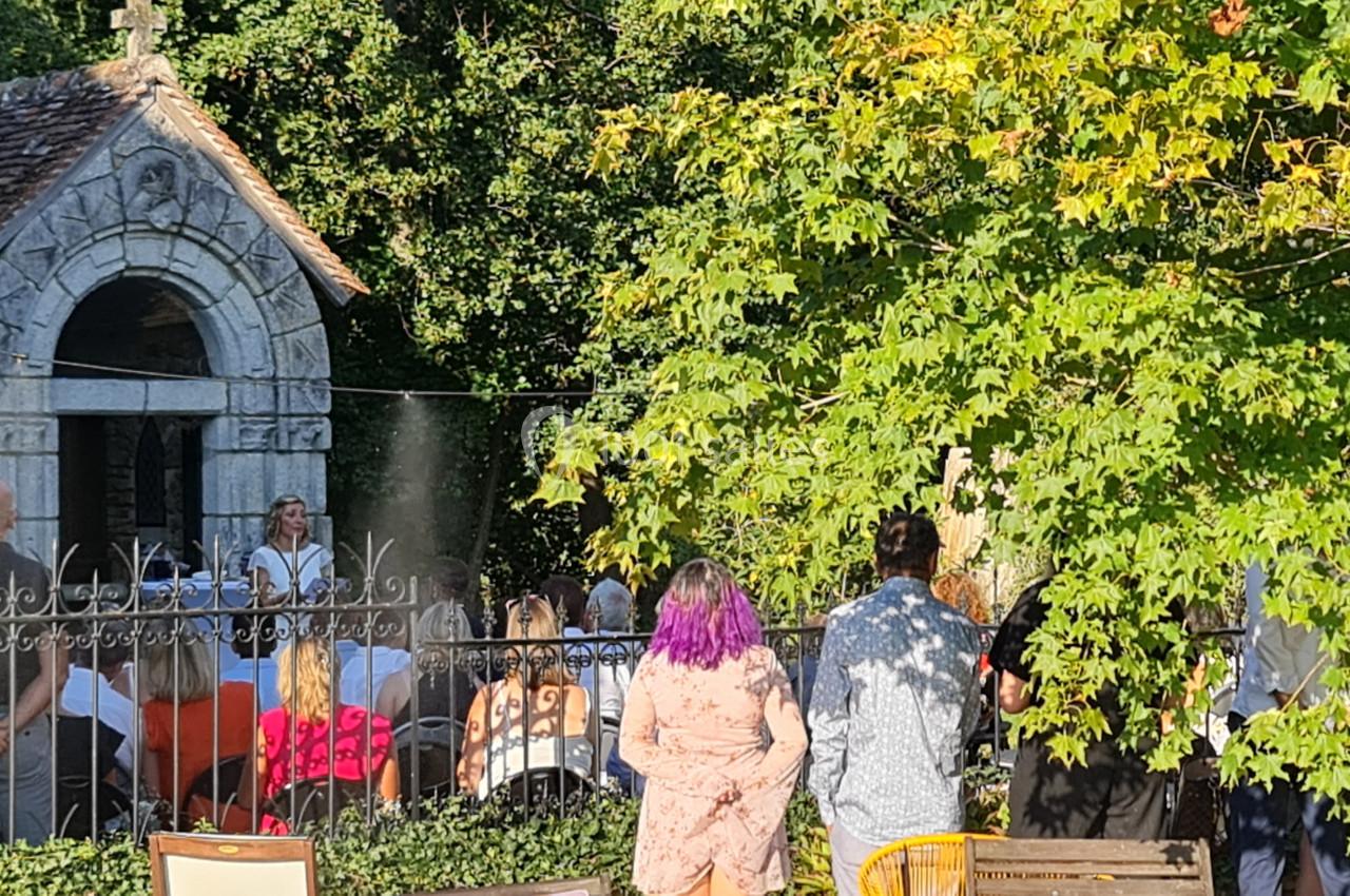Un groupe de personnes assiste à un événement en plein air près d'une petite chapelle en pierre entourée de verdure.