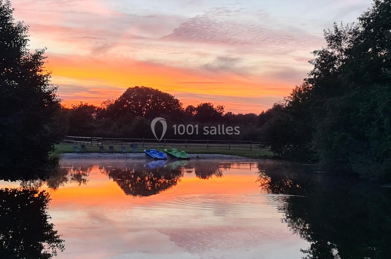 Étang calme au coucher du soleil, avec des arbres, des barques colorées sur la rive et des reflets dans l'eau.