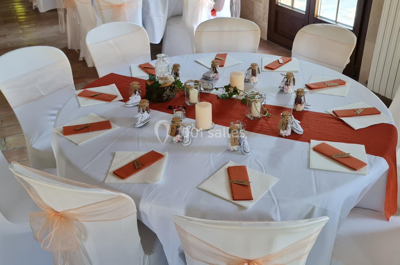Table ronde décorée pour un événement, avec nappes blanches, chemin de table orange, bougies et petits ornements.