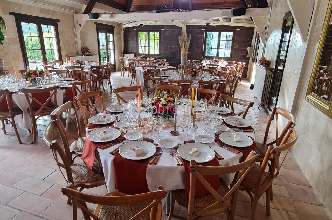 Salle de réception lumineuse avec tables rondes dressées, décorées de fleurs colorées et serviettes rouges.