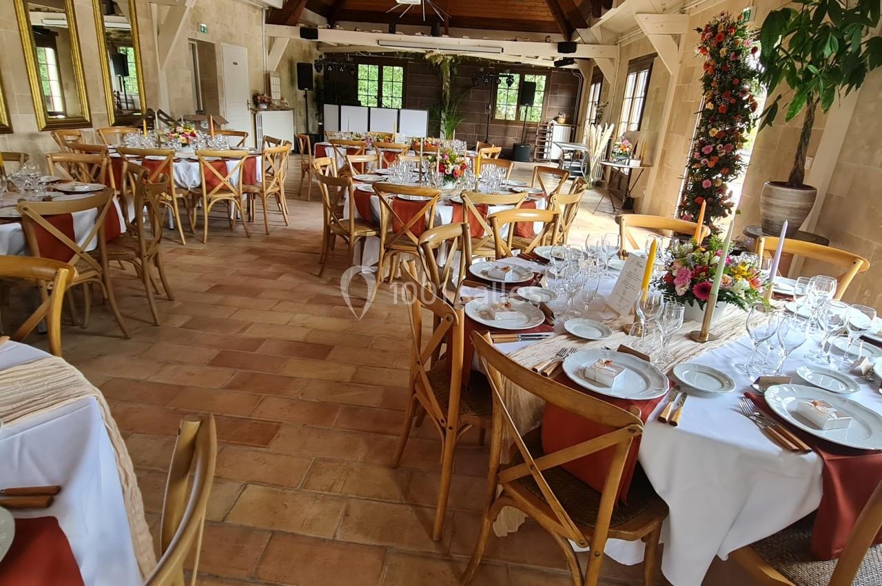 Salle de réception décorée avec des tables rondes dressées, nappes blanches, chaises en bois et compositions florales.