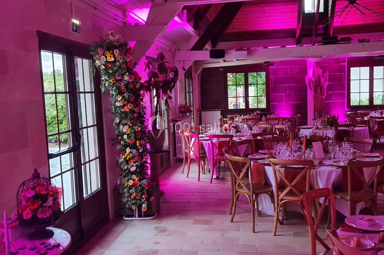 Salle de réception décorée avec des tables dressées, des fleurs et un éclairage rose.