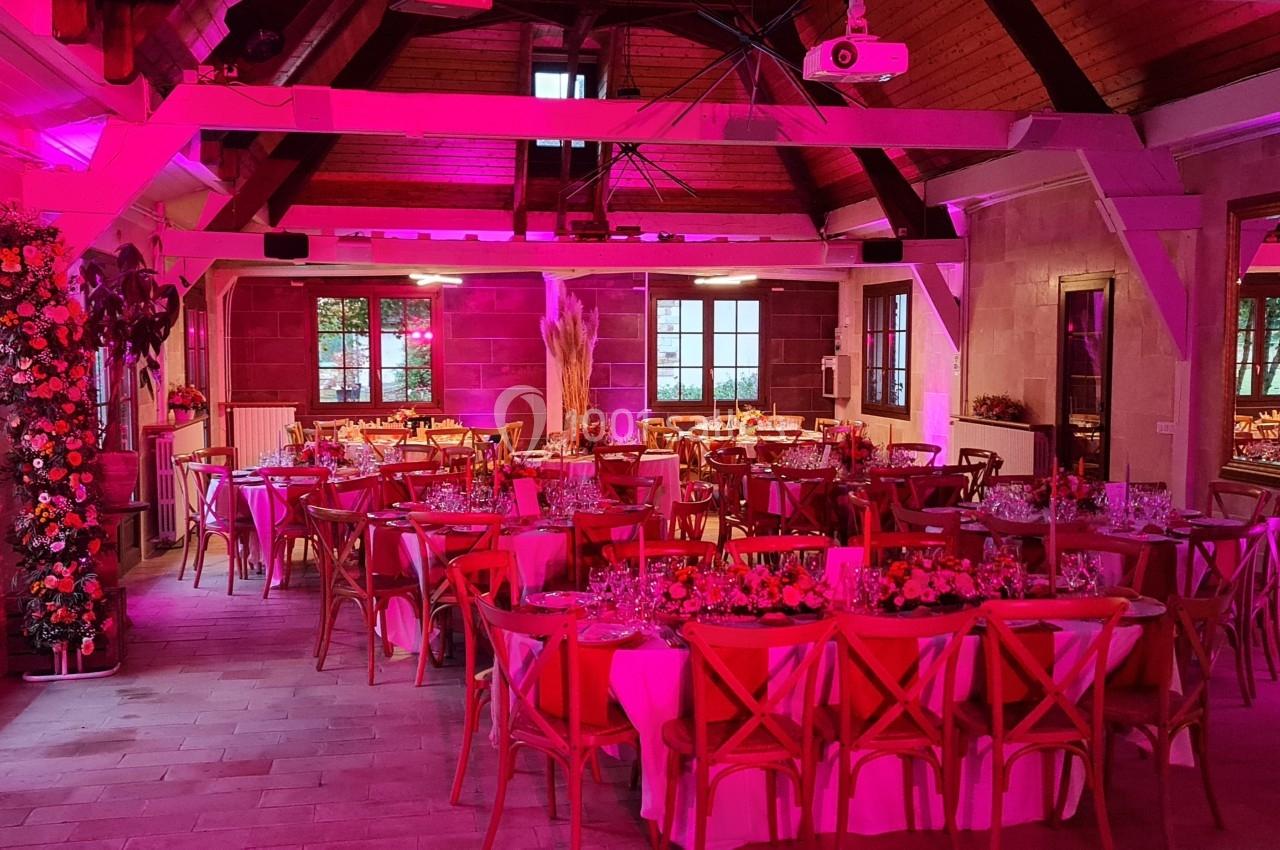 Salle de réception décorée avec des tables rondes, des chaises en bois et un éclairage rose.
