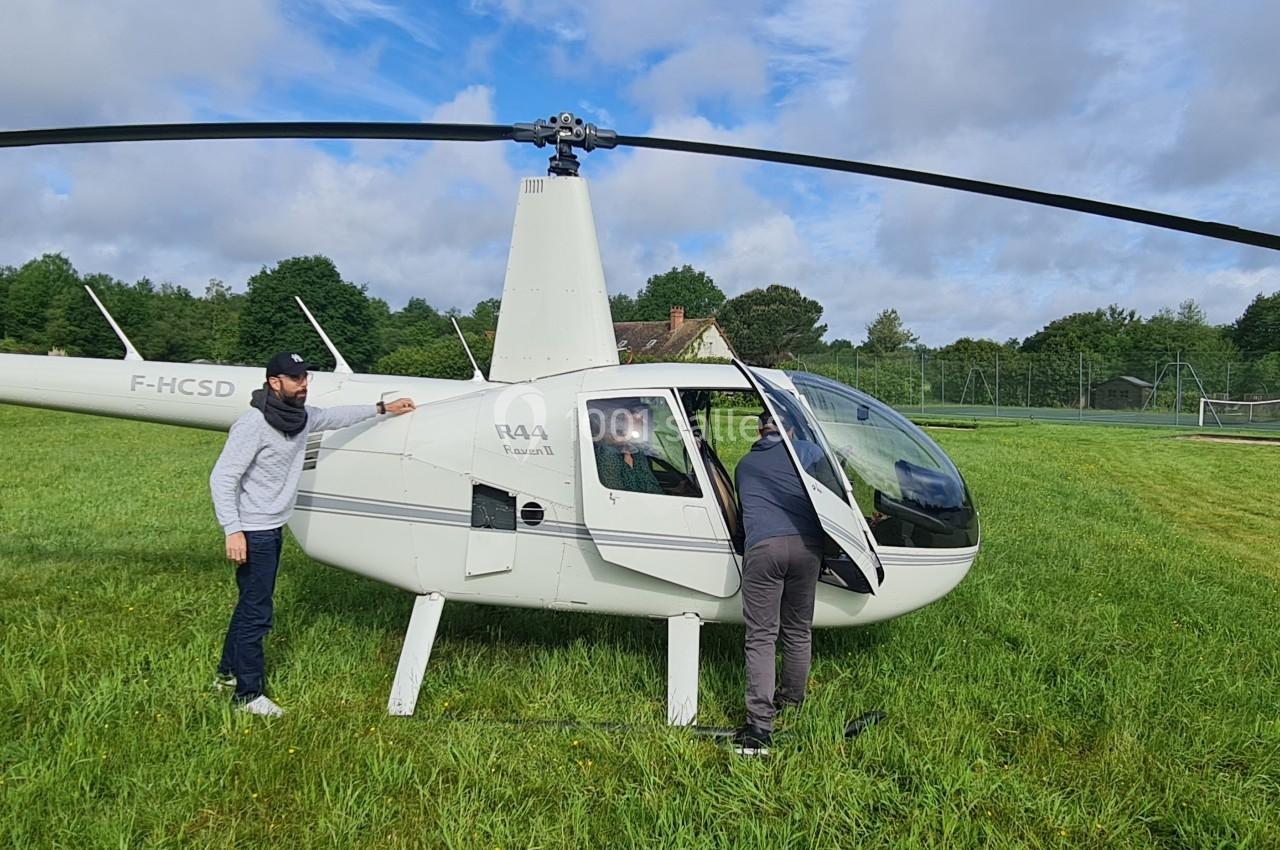 Deux hommes inspectent un petit hélicoptère blanc stationné sur une pelouse, entouré de verdure et de ciel nuageux.