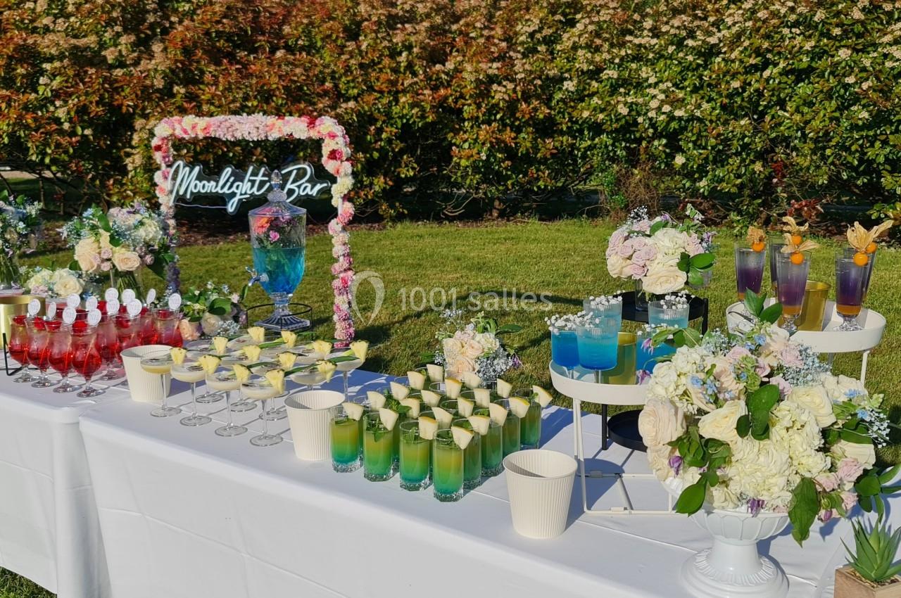 Table de cocktails colorés et décorations florales disposée en extérieur, devant un panneau ’Moonlight Bar’.