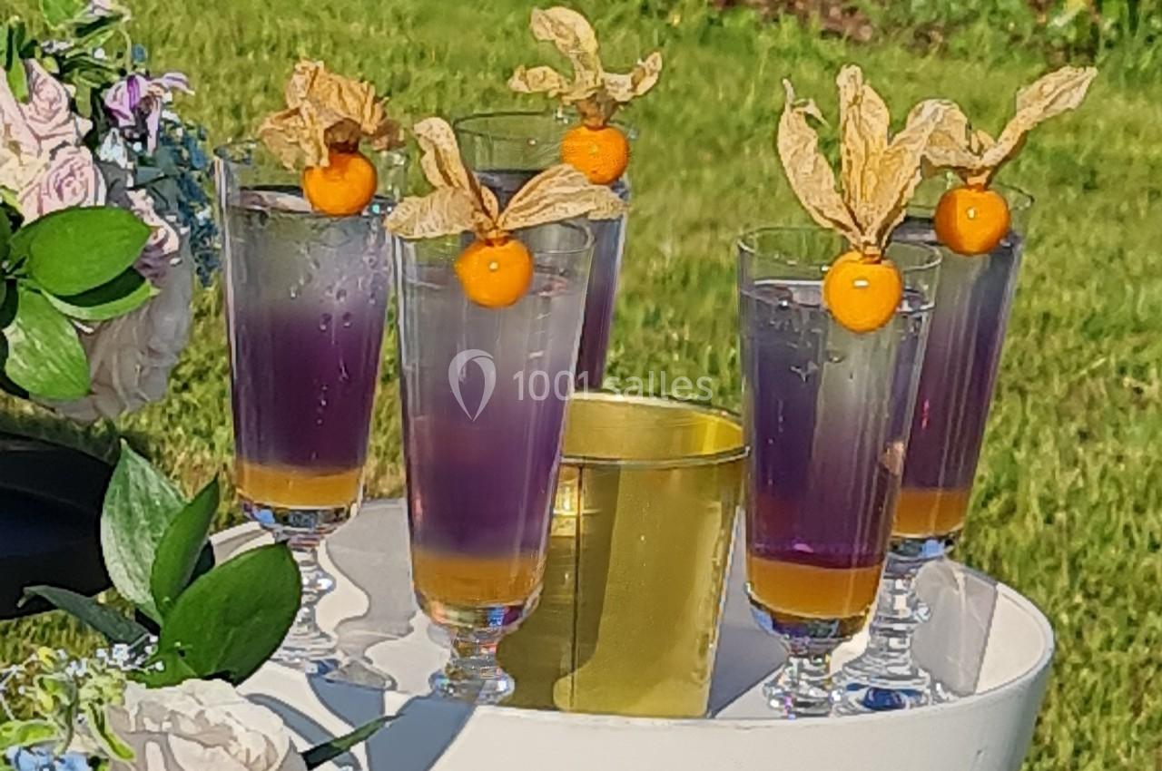 Verres de cocktail violets garnis de physalis, posés sur une table blanche décorée de fleurs, en extérieur.