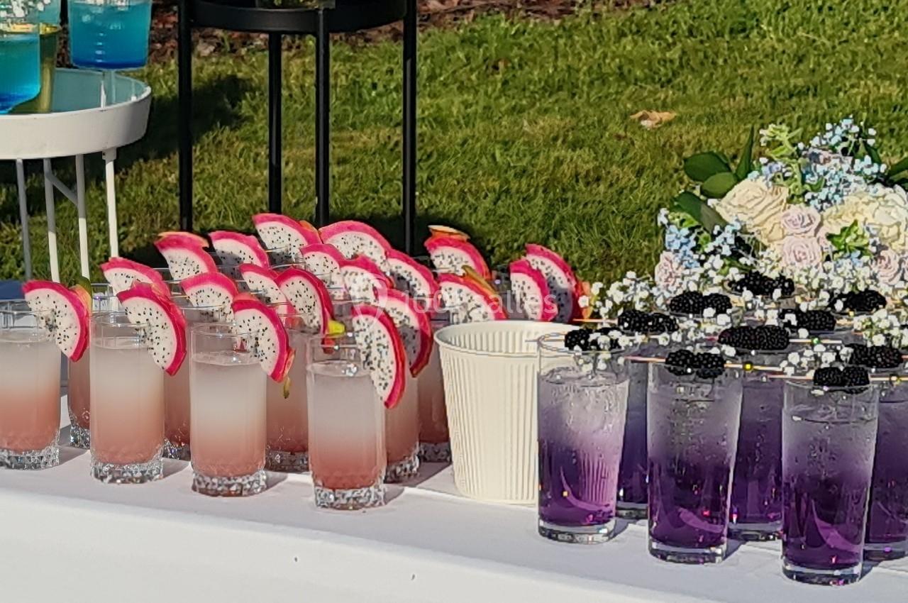 Table avec des boissons colorées, dont des cocktails roses à la pitaya, violets avec mûres et verts, décorée de fleurs.