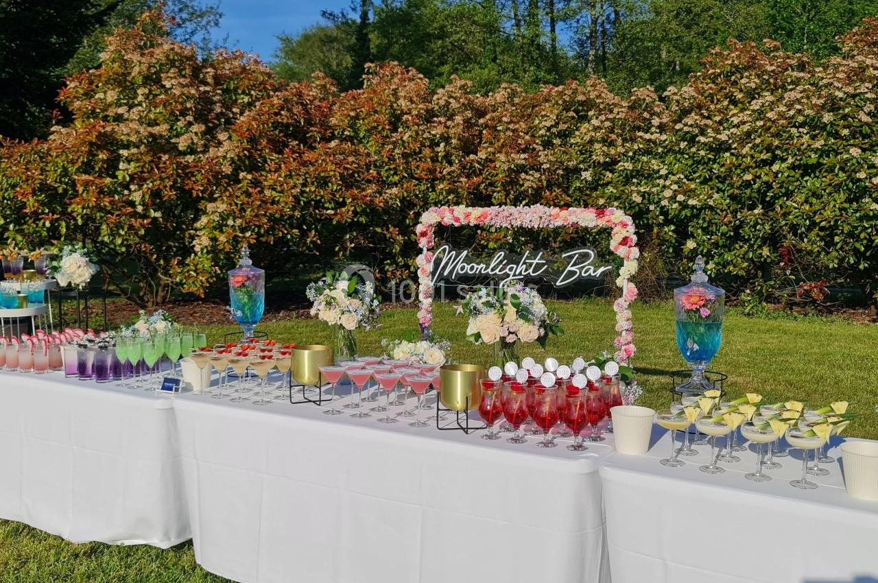 Table extérieure décorée avec des boissons colorées, fleurs et un panneau lumineux ’Moonlight Bar’, devant des arbustes…