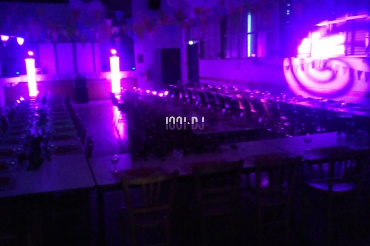Salle sombre avec tables et chaises alignées, éclairée par des lumières violettes et des projecteurs lumineux.