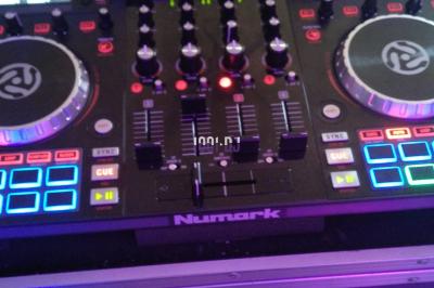 Table de mixage DJ Numark avec deux platines, des boutons lumineux et un écran, posée sur un support.